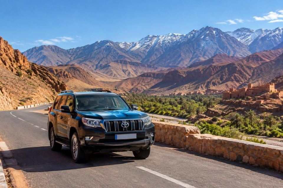 A private 4x4 with a driver on a High Atlas mountain road, snow-capped peaks behind and a kasbah in the palm valley below.